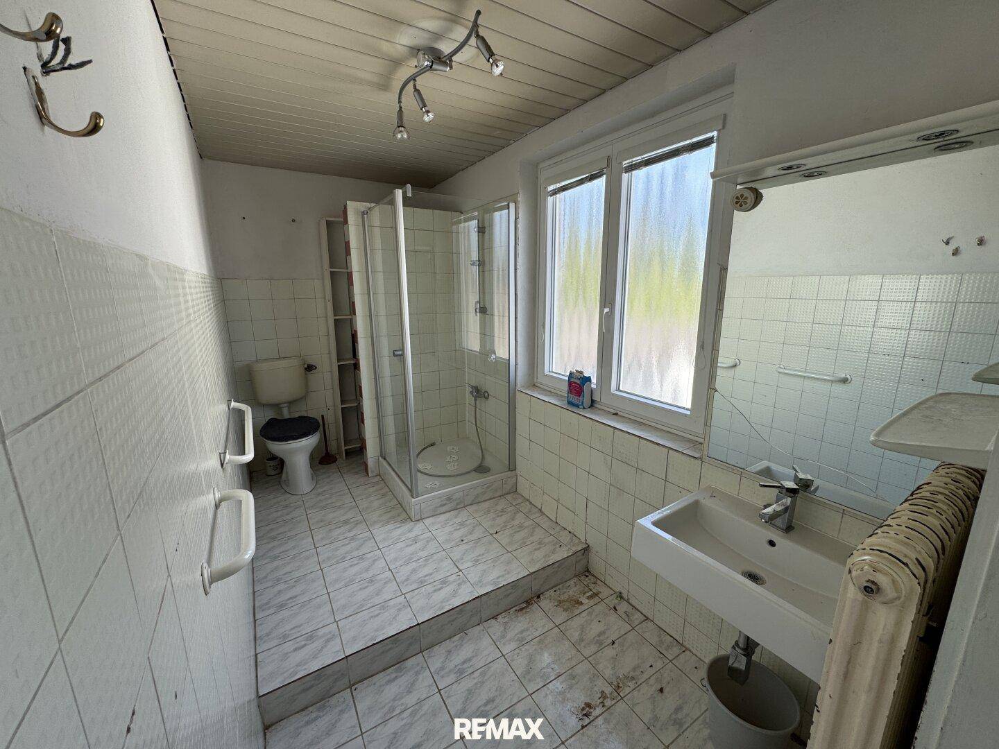 Badezimmer mit Duschkabine, Toilette, Waschbecken und Fenster, ausgestattet mit Haltegriffen.