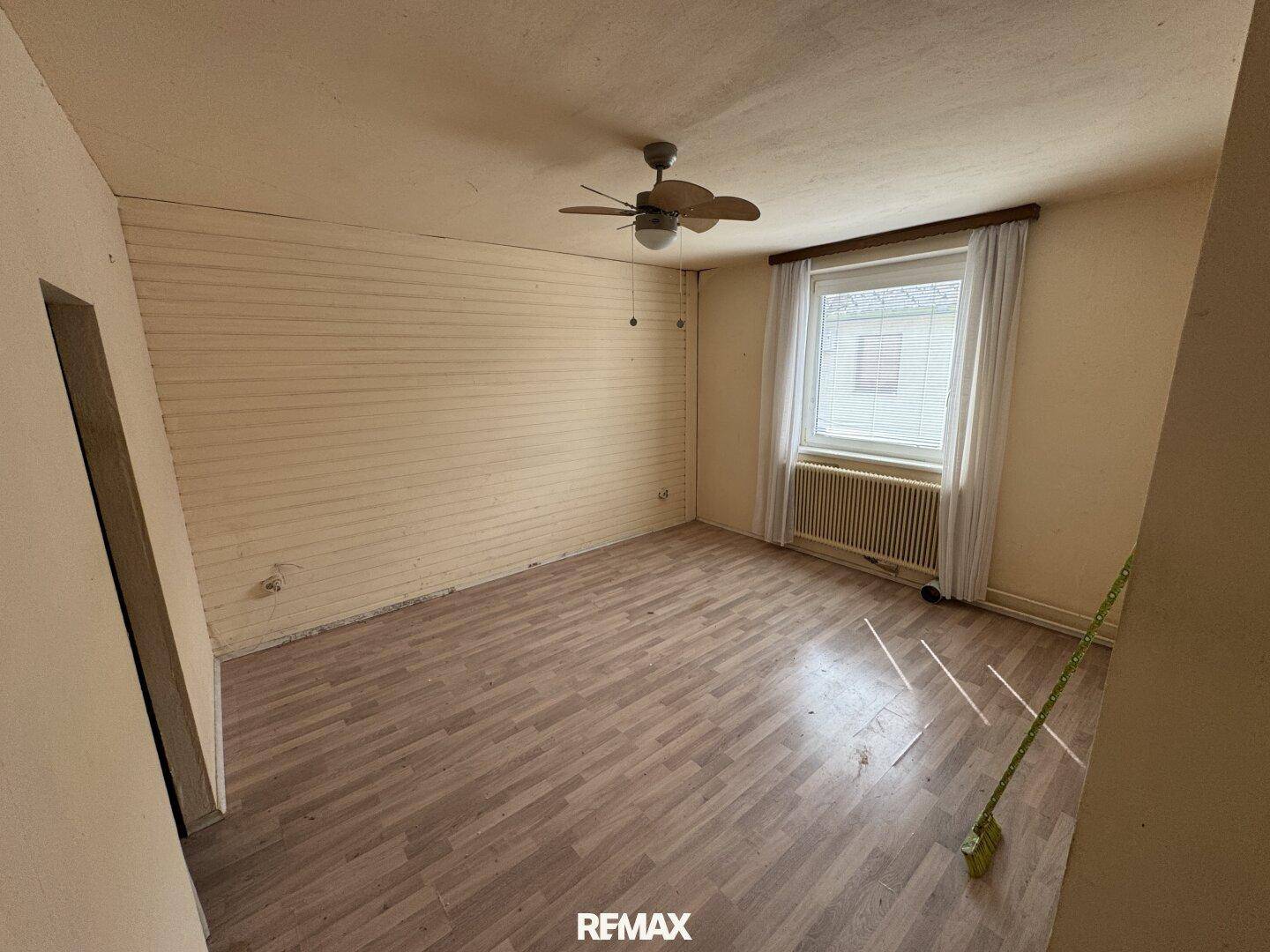 Leeres Zimmer mit Holzparkettboden, Fenster und Deckenventilator, in renovierungsbedürftigem Zustand.