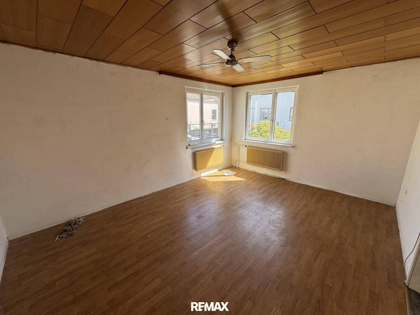 Leeres Zimmer mit Holzdecke, Laminatboden und zwei Fenstern, bereit zur Renovierung.