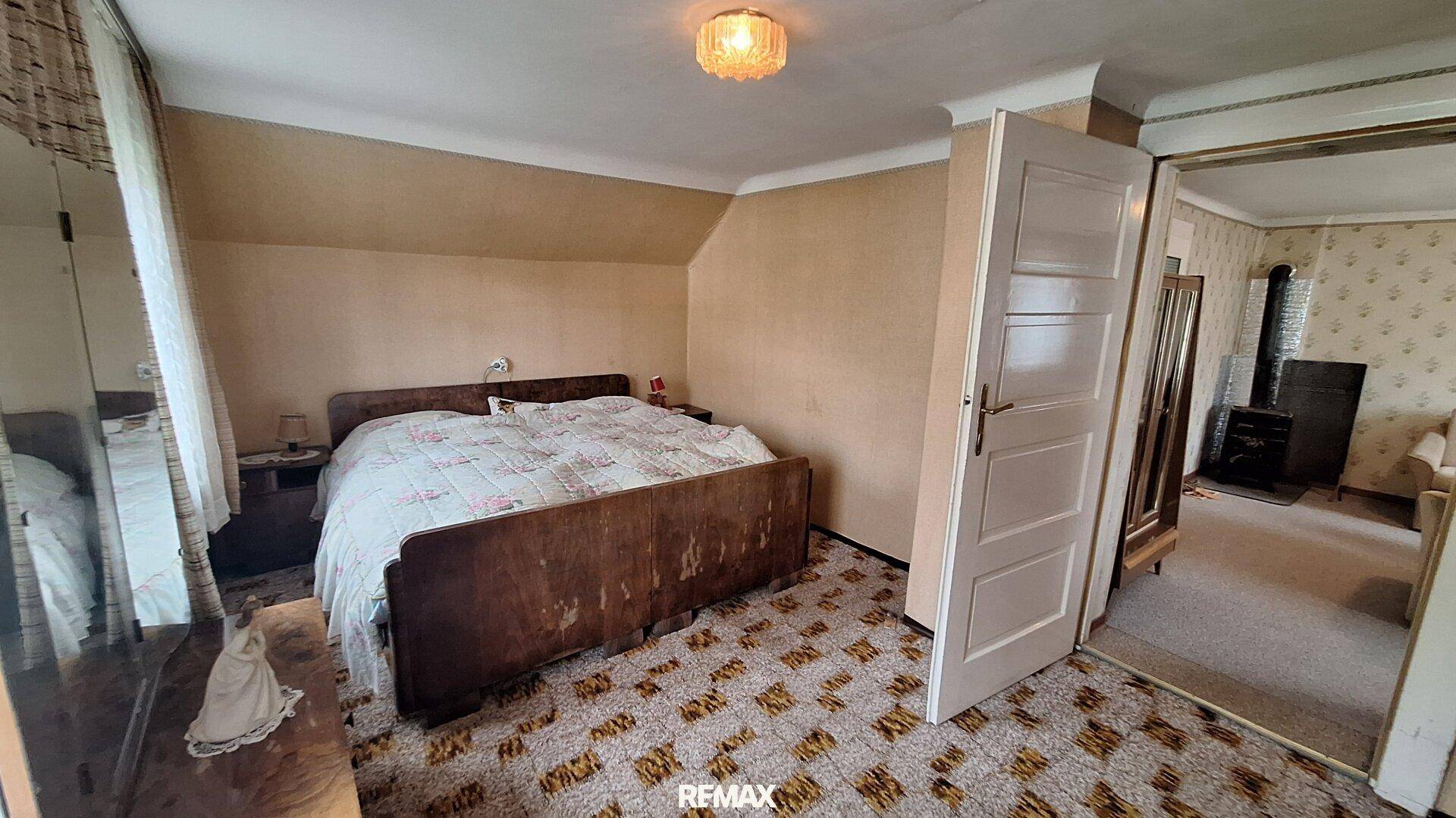 Schlafzimmer mit schrägem Dach, älterem Holzbett und gemustertem Teppichboden.