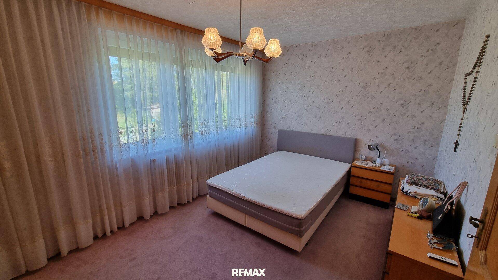 Schlafzimmer mit Doppelbett, rosa Teppichboden und großen Fenstern mit Vorhängen.