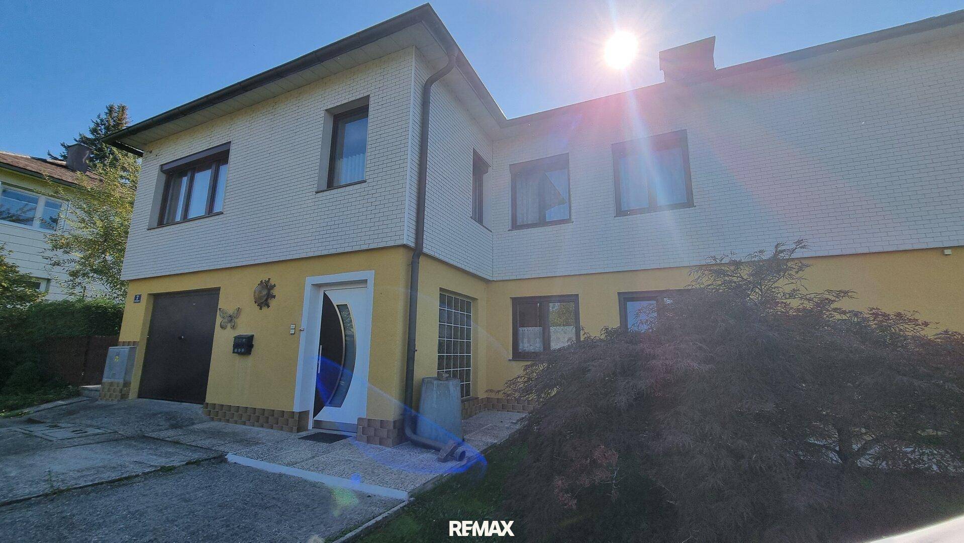 Vorderansicht des Hauses mit gelb-weißer Fassade, Garage und Eingangsbereich unter strahlendem Sonnenschein.