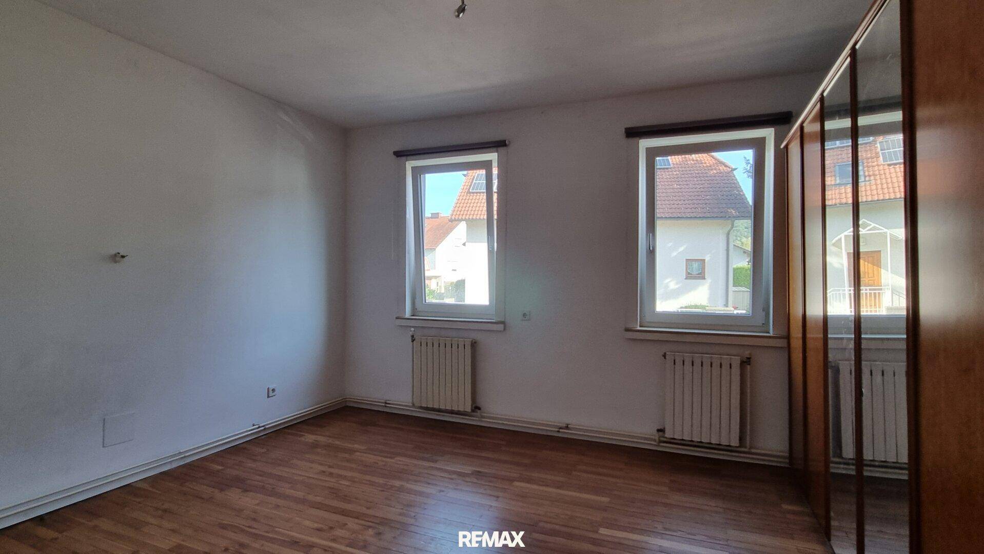 Helles Schlafzimmer mit Holzboden, zwei Fenstern und einem Spiegelschrank.