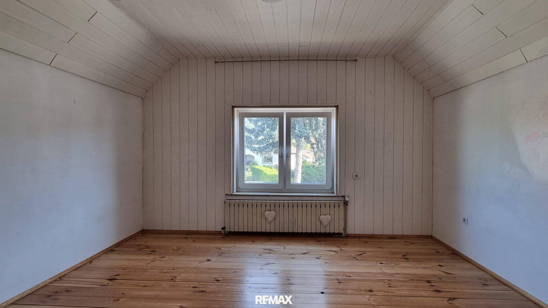 Helles Zimmer im Dachgeschoss mit Holzdielenboden, schrägen Wänden und einem Fenster.