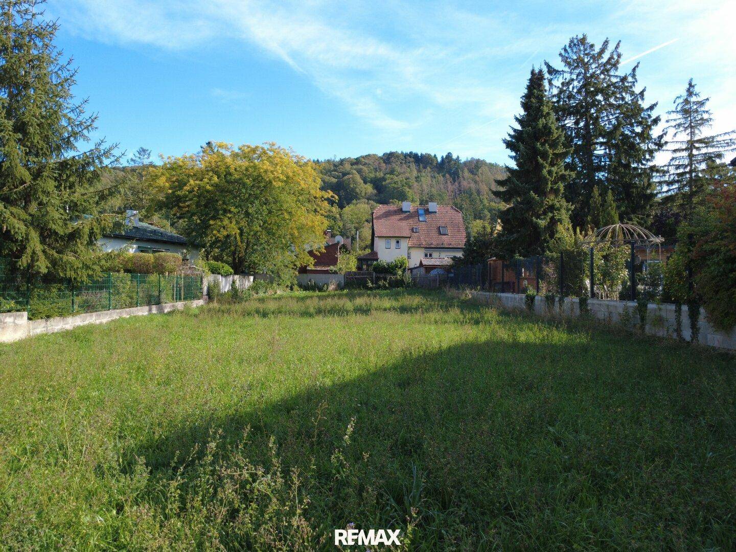 Blick über das grüne Grundstück mit umliegenden Häusern und bewaldeten Hügeln im Hintergrund.