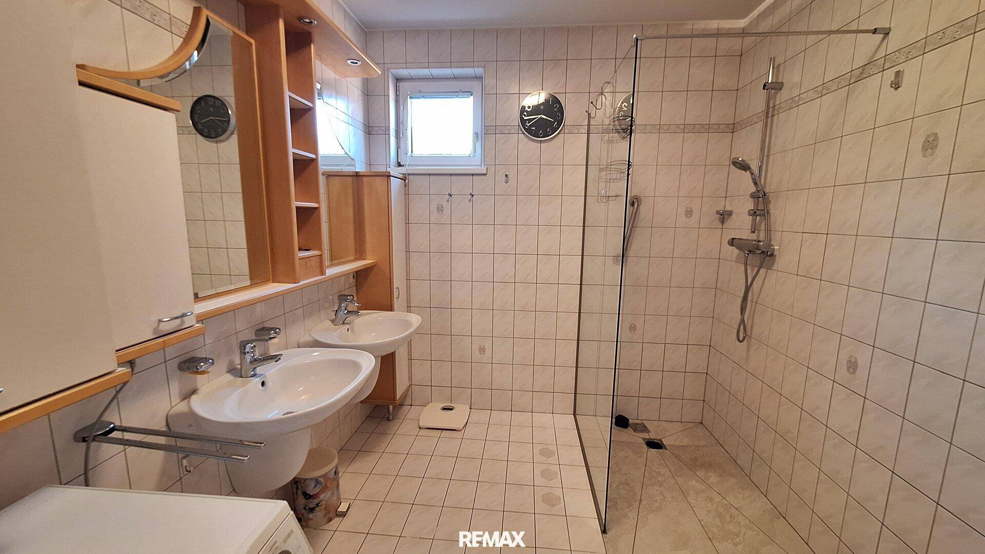 Gefliestes Badezimmer mit Doppelwaschbecken, Dusche und Fenster.