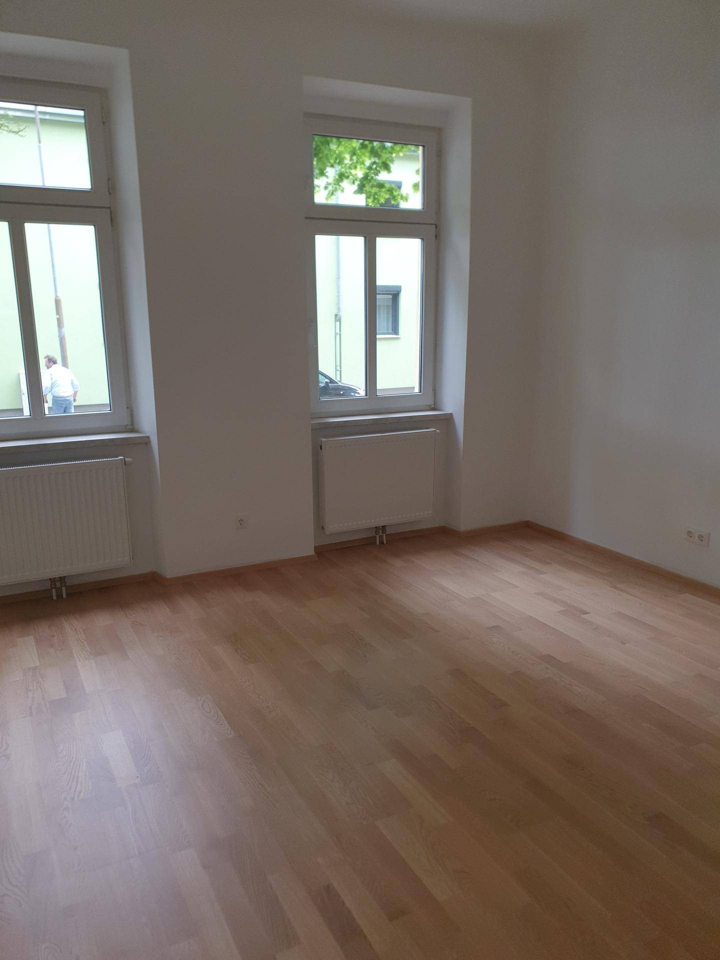 Freundliches Zimmer mit hellem Holzparkett, weißen Wänden und zwei großen Fenstern.