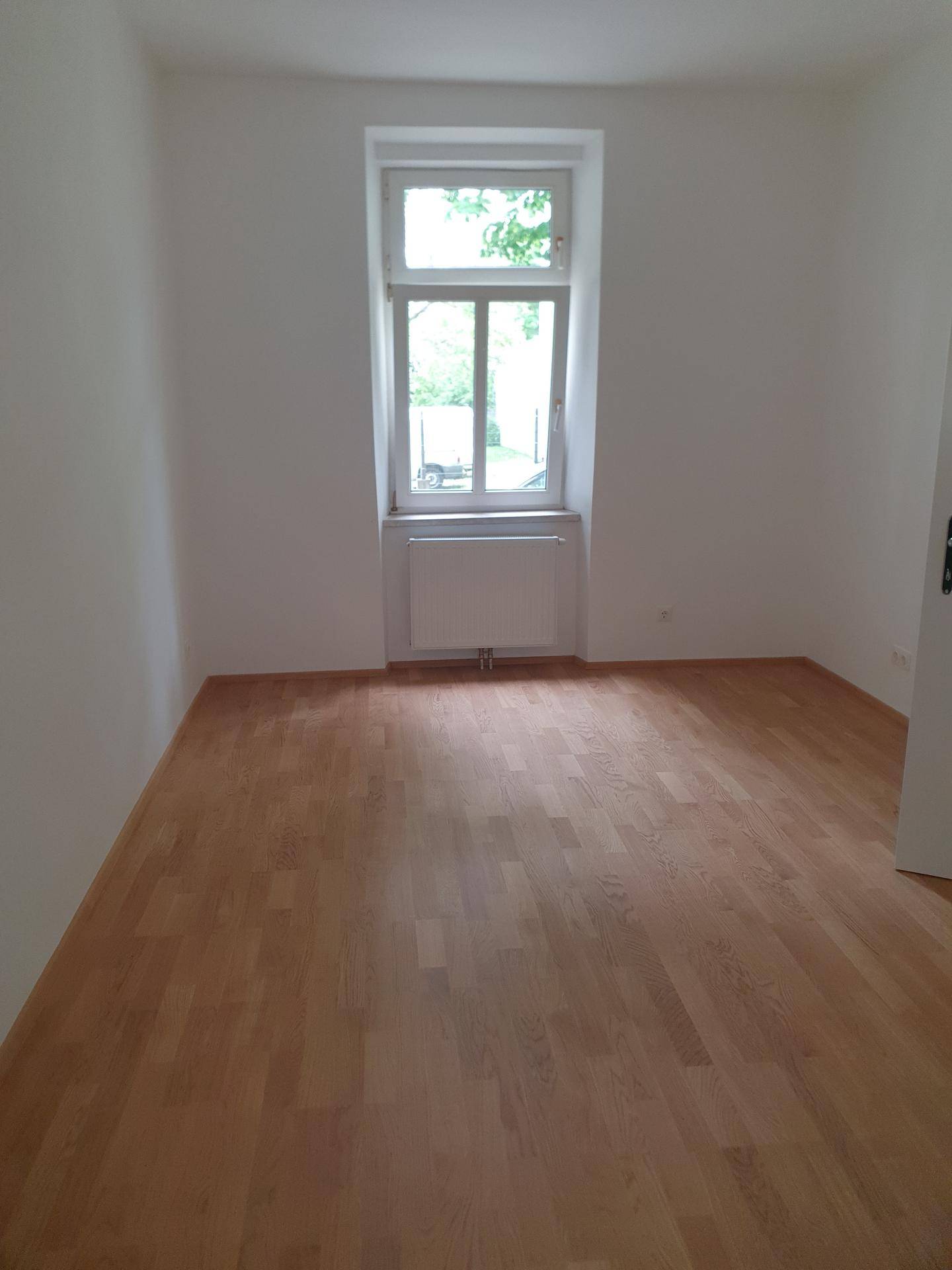 Freundliches Zimmer mit hellem Holzparkett, weißen Wänden und einem großen Fenster.