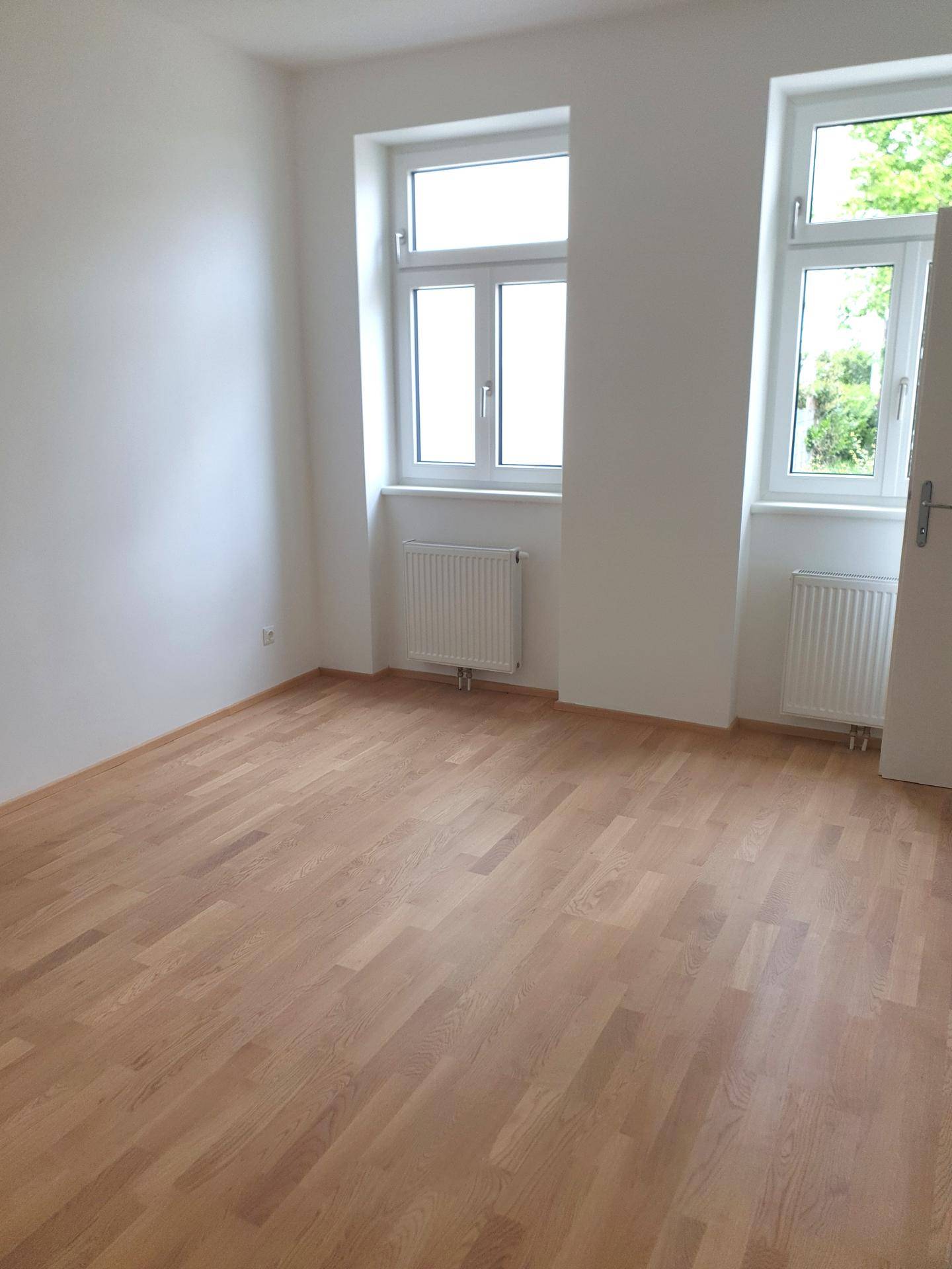 Helles Zimmer mit neuem Holzparkettboden, weißen Wänden und zwei Fenstern mit Heizkörpern.