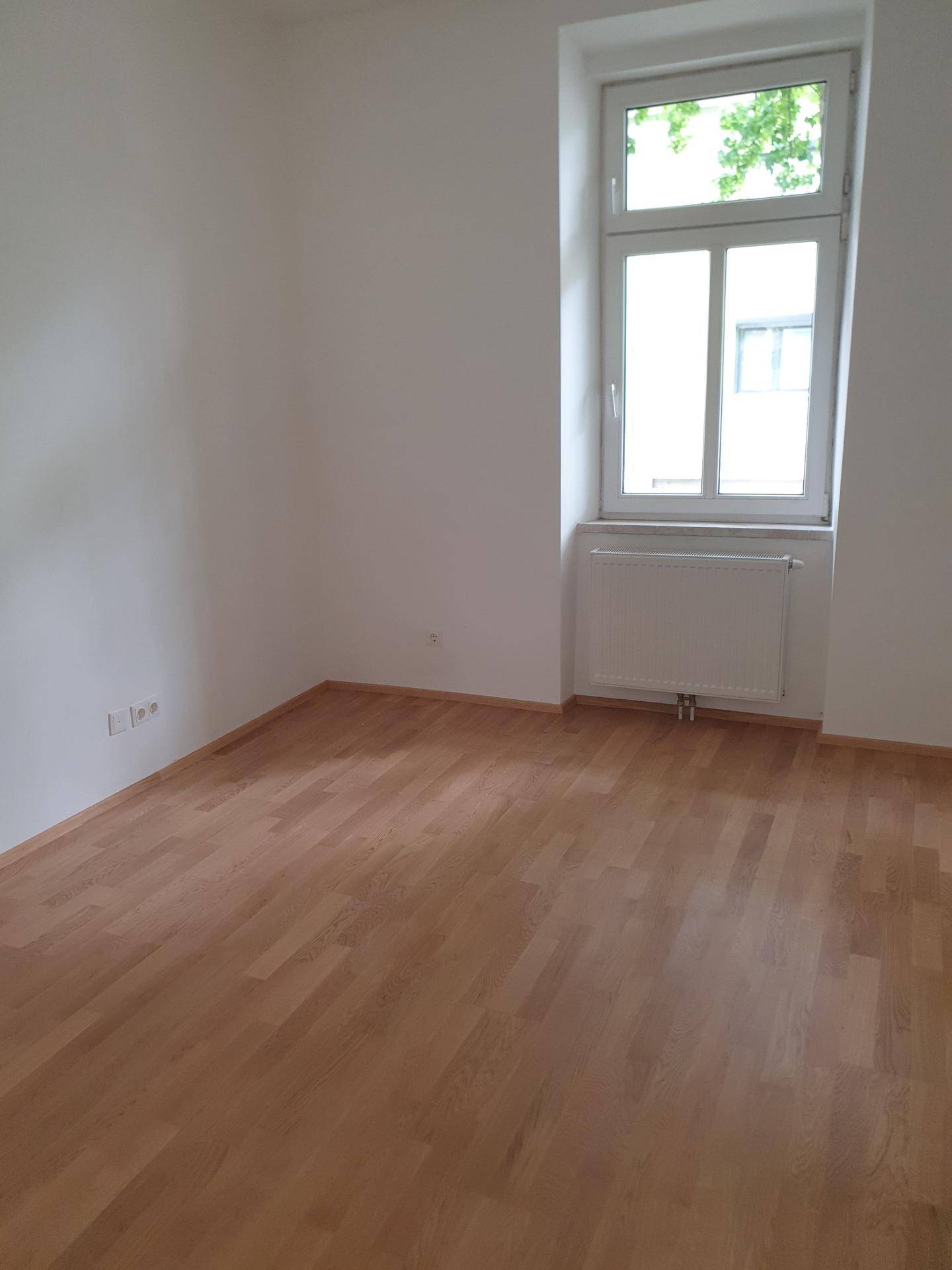 Helles Zimmer mit neuem Holzparkettboden, weißen Wänden und einem Fenster mit Heizkörper.