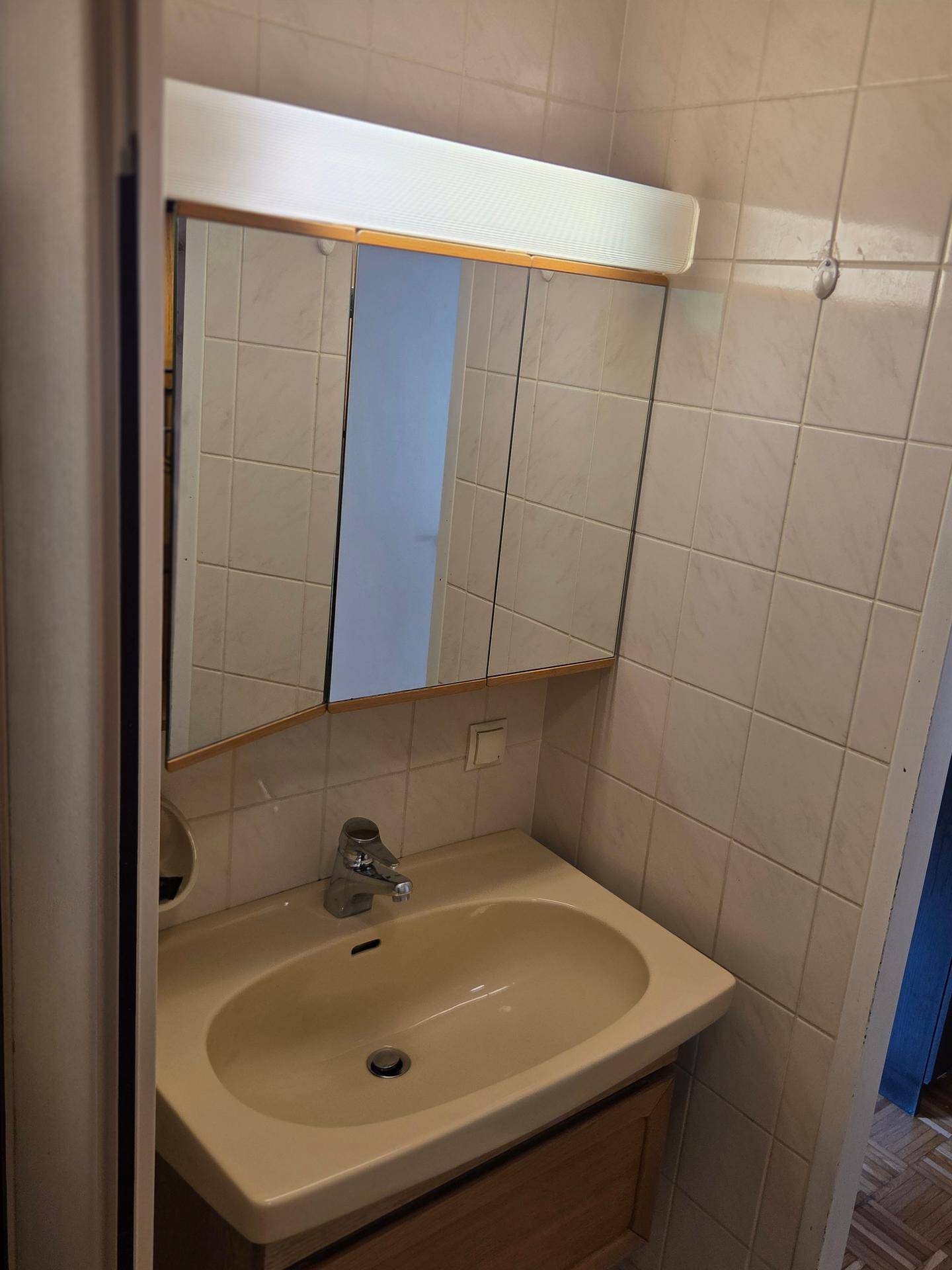 Badezimmer mit Waschbecken und Spiegelschrank darüber, umgeben von hellen Fliesen.