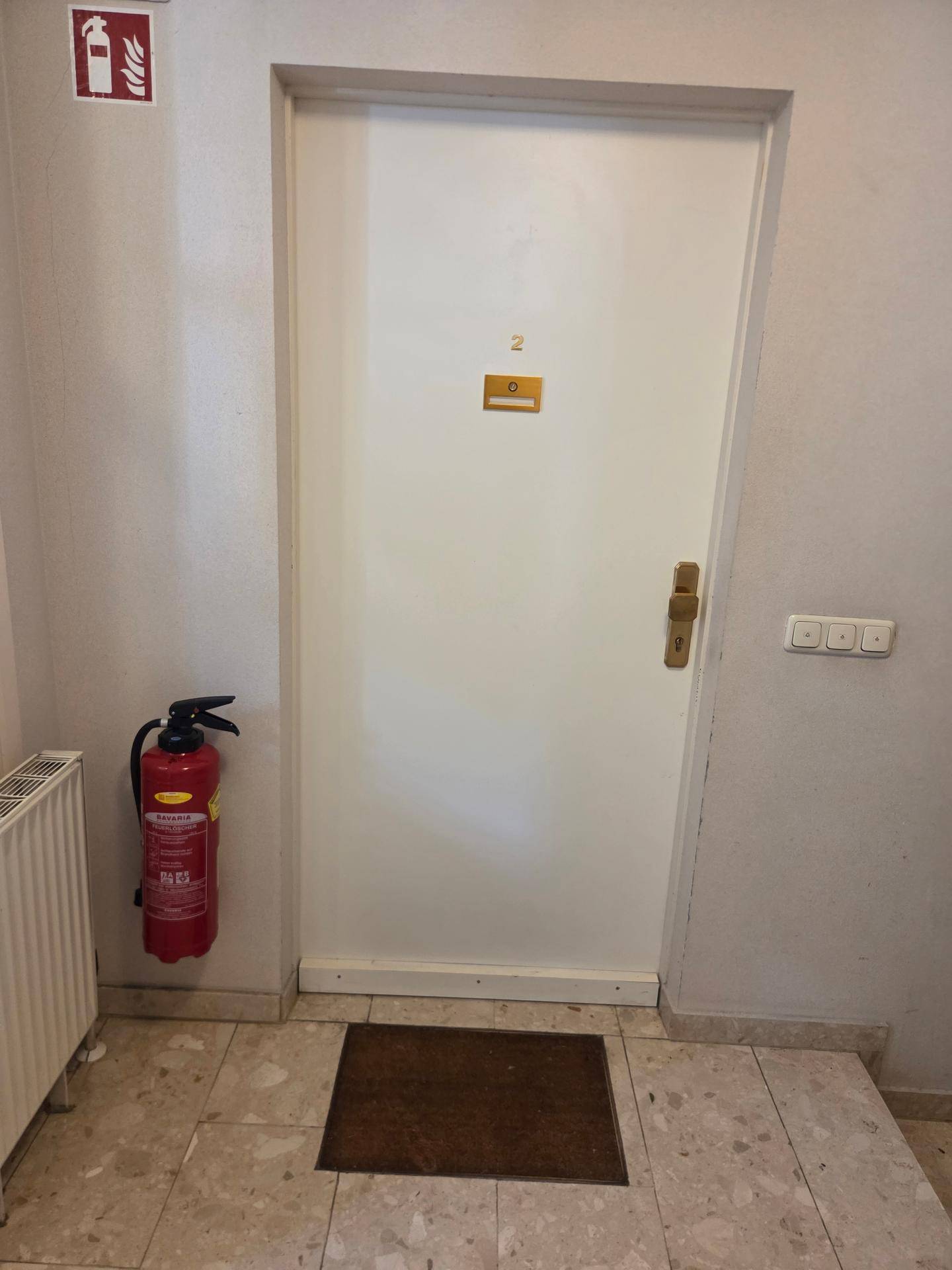 Heller Flur mit weißer Tür, Fliesenboden und einem Feuerlöscher an der Wand.