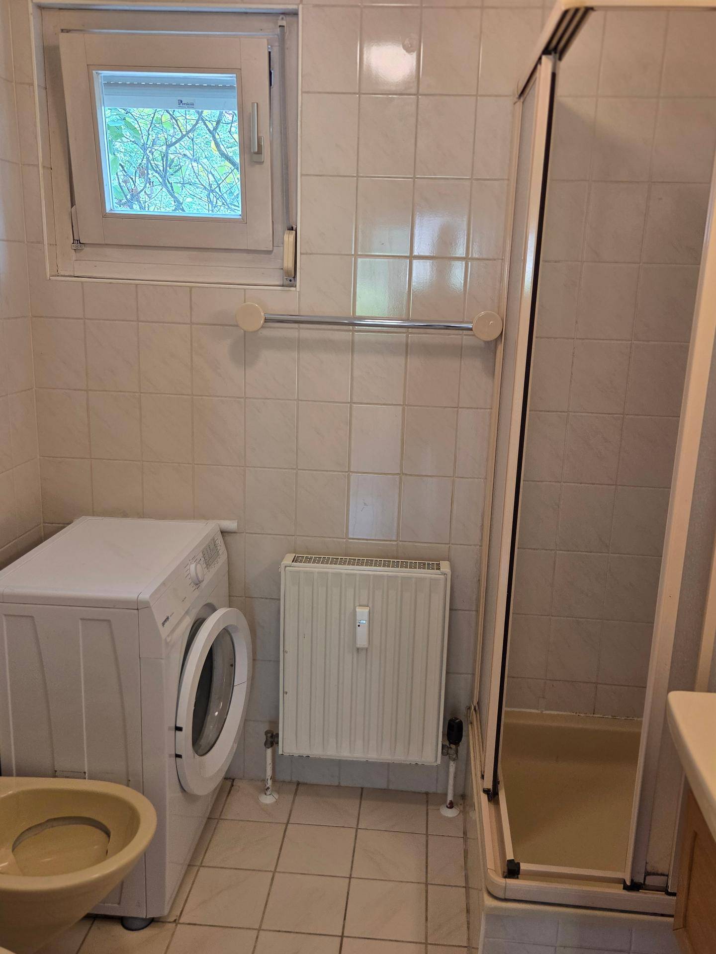 Gefliestes Badezimmer mit Duschkabine, Waschmaschine und kleinem Fenster für Tageslicht.