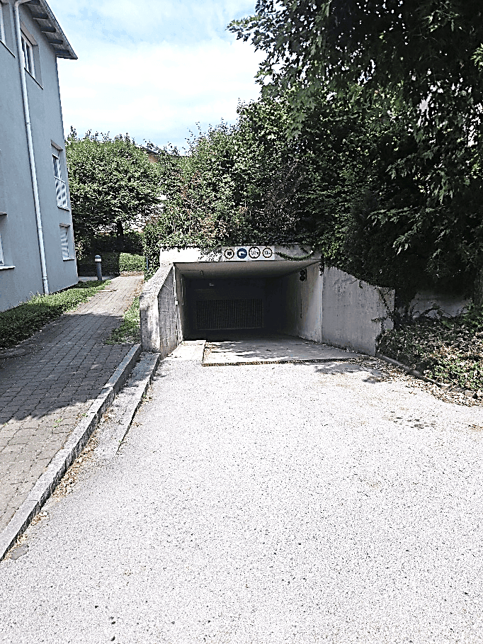Einfahrt zur Tiefgarage von der Straße aus, umgeben von Bäumen und Büschen.
