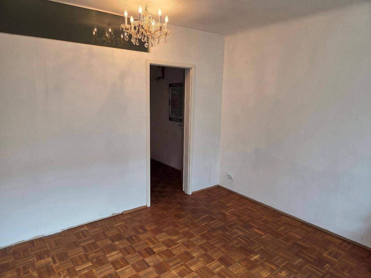 Geräumiges Zimmer mit Parkettboden, Kronleuchter und einem Durchgang zu einem weiteren Raum.