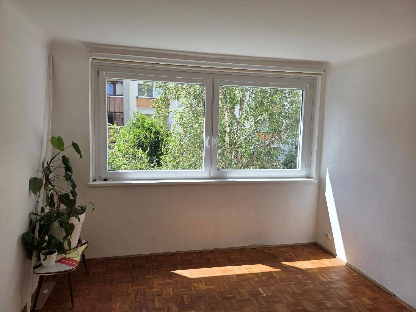 Helles Zimmer mit Parkettboden und großem Fenster, das einen Blick ins Grüne bietet.