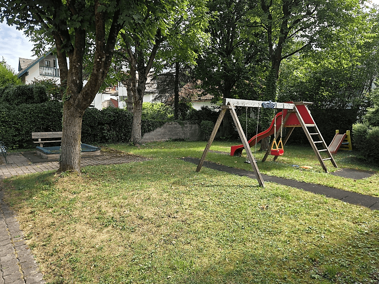 Spielplatz im Freien mit Schaukeln, Rutsche und Sandkasten, umgeben von Bäumen und Rasen.