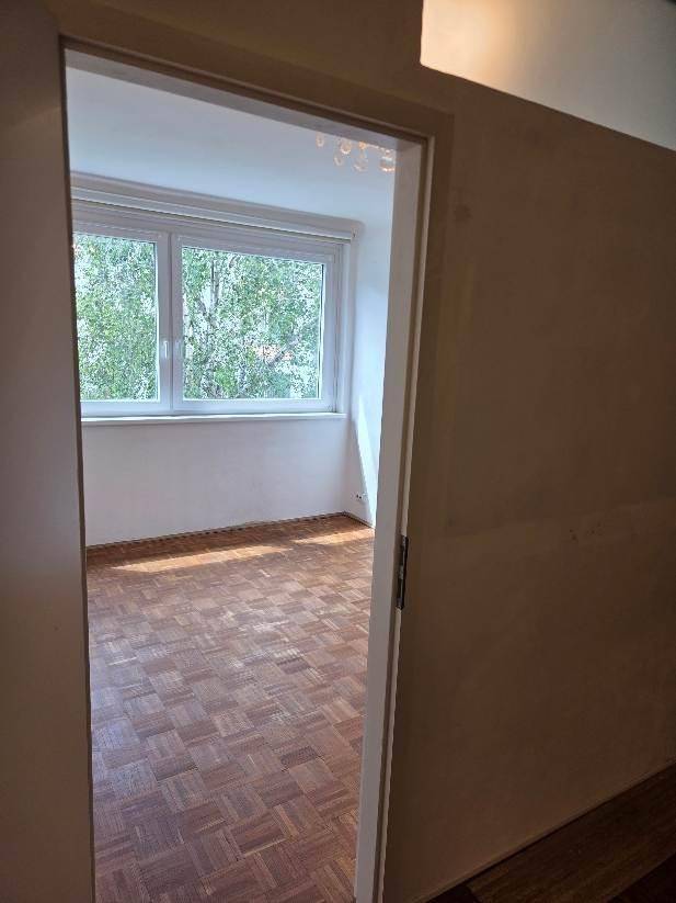 Blick durch eine Tür in ein helles Zimmer mit Parkettboden und großem Fenster mit Grünblick.
