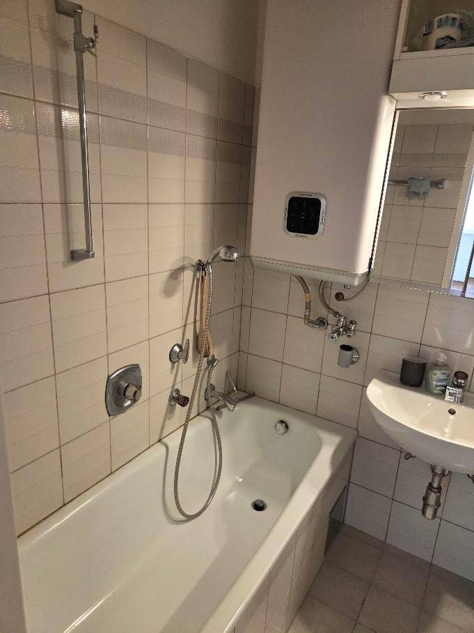 Badezimmer mit weißen Fliesen, Badewanne mit Duschfunktion, Waschbecken und Warmwasserbereiter.