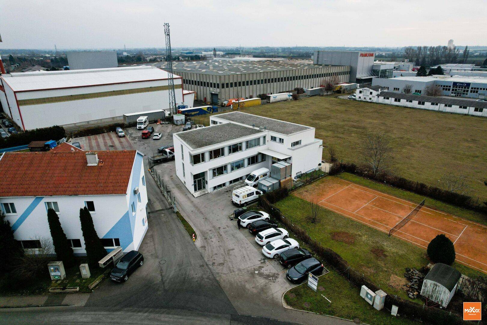 Luftaufnahme eines modernen Bürogebäudes umgeben von Industrieanlagen, einem Tennisplatz und Grünflächen.