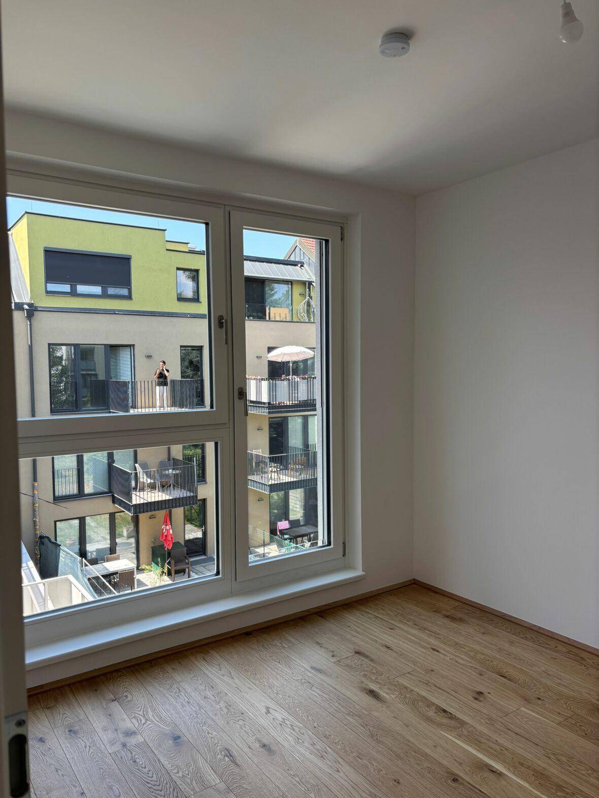 Helles Zimmer mit Holzboden und Blick auf den Innenhof mit weiteren Balkonen.