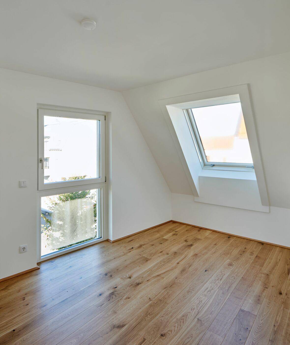 Helles Zimmer im Dachgeschoss mit Holzboden, Dachfenster und einem weiteren Fenster.