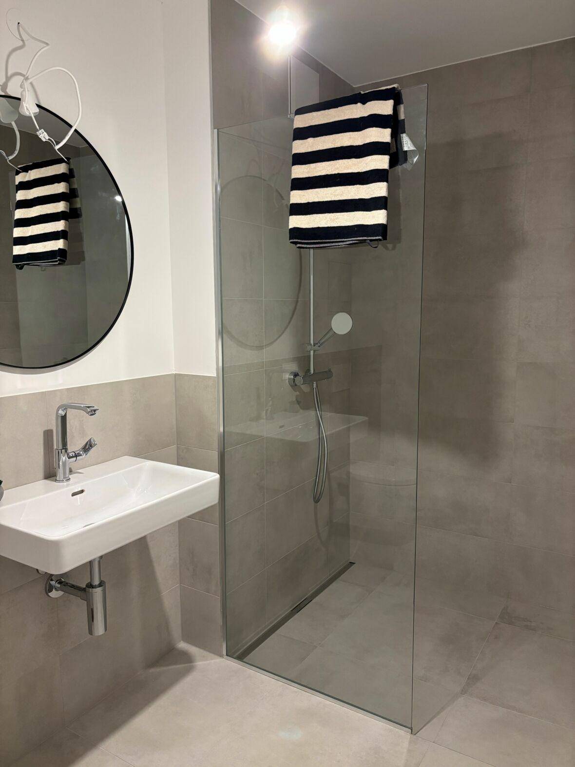Modernes Badezimmer mit grauen Fliesen, wandmontiertem Waschbecken und einer begehbaren Dusche mit Glasabtrennung.