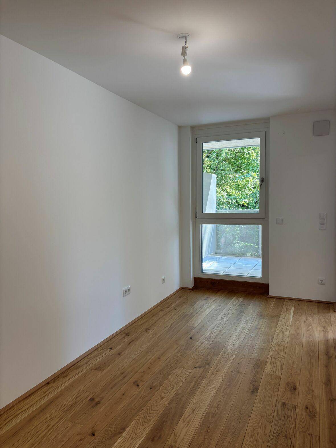 Lichtdurchfluteter Raum mit Holzboden, weißen Wänden und einem großen Fenster.