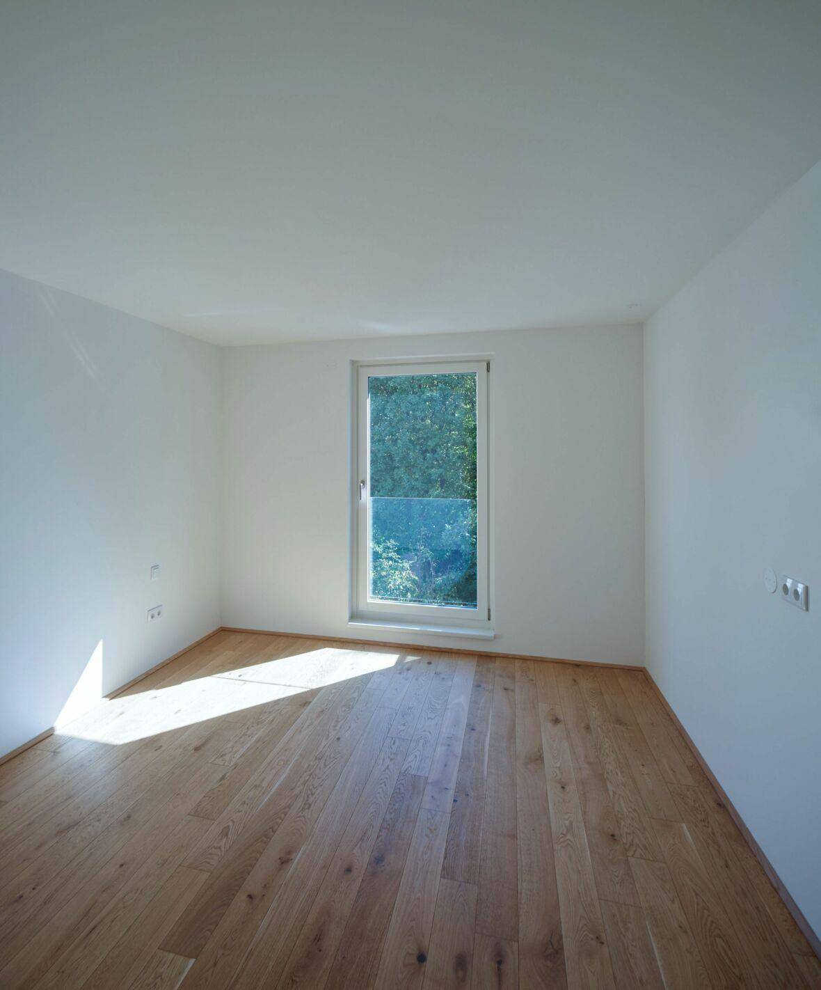 Helles, leeres Zimmer mit Holzboden und Fenster mit Blick ins Grüne, ideal als Schlafzimmer.