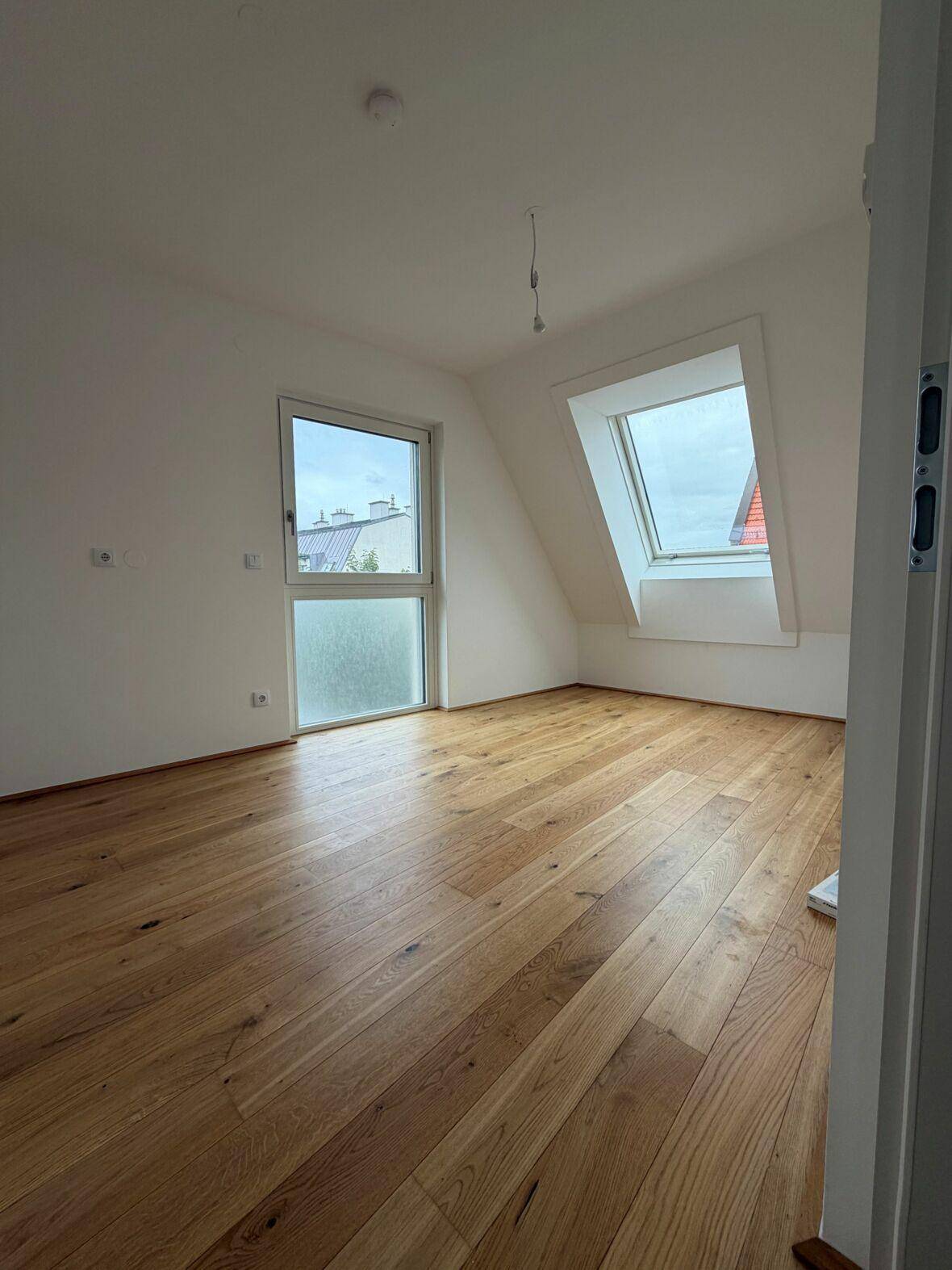 Helles Zimmer im Dachgeschoss mit Echtholzboden, Dachschrägen und zwei Fenstern, darunter ein großes Dachfenster.
