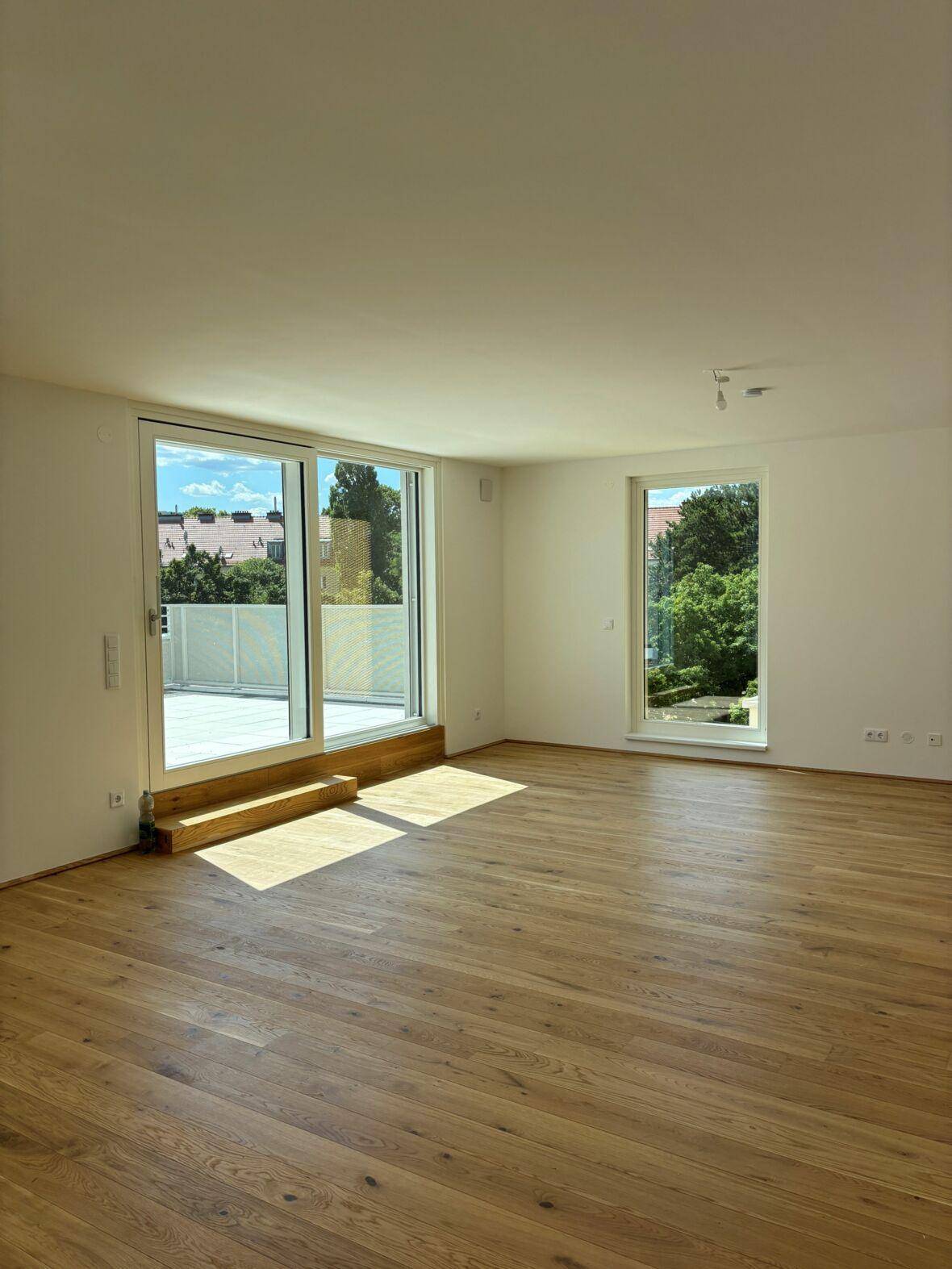Helles, leeres Wohnzimmer mit Holzboden und Zugang zu einer Terrasse, durchflutet von Sonnenlicht.