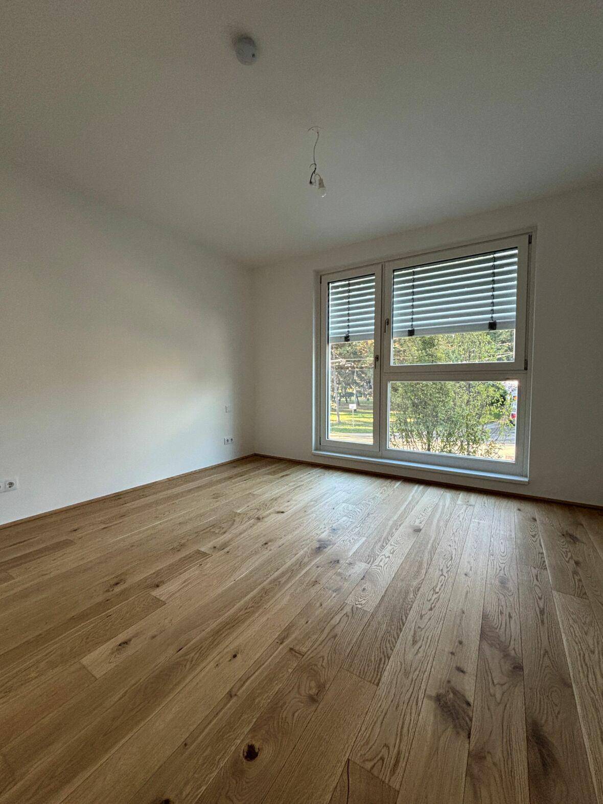 Helles Schlafzimmer mit Parkettboden und einem Fenster mit Jalousien, das einen Blick ins Grüne bietet.
