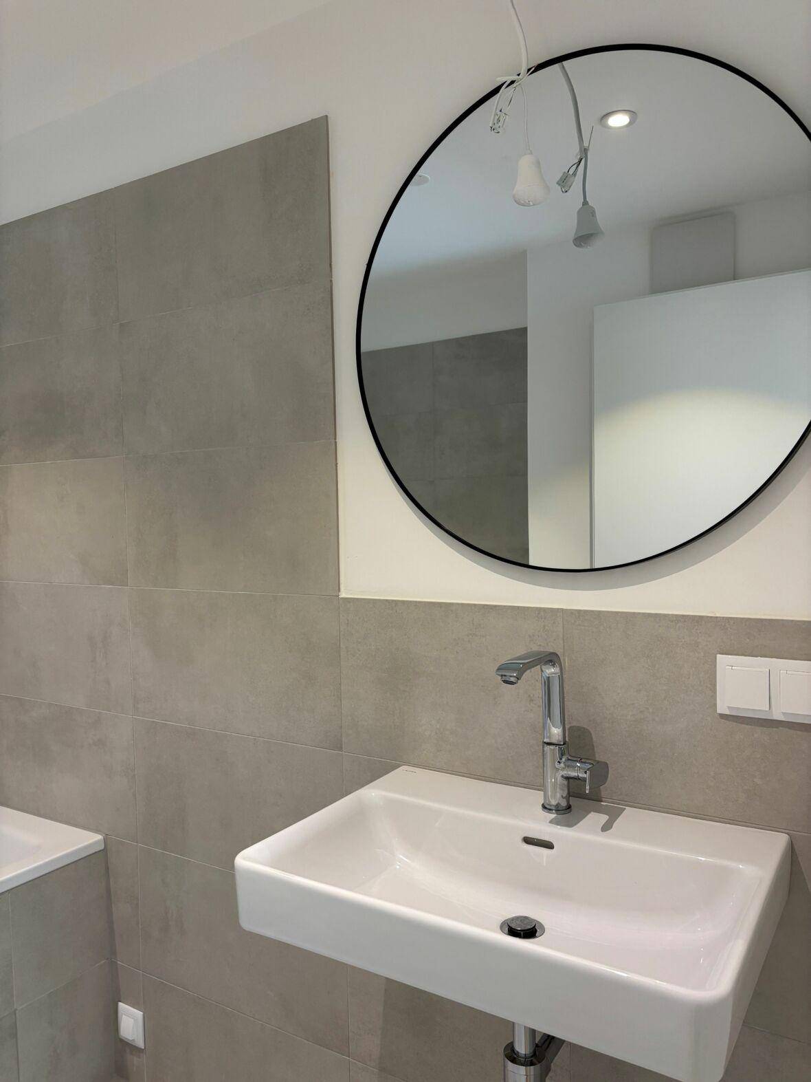 Modernes Badezimmerdetail mit wandmontiertem Waschbecken und einem runden Spiegel über grauen Fliesen.
