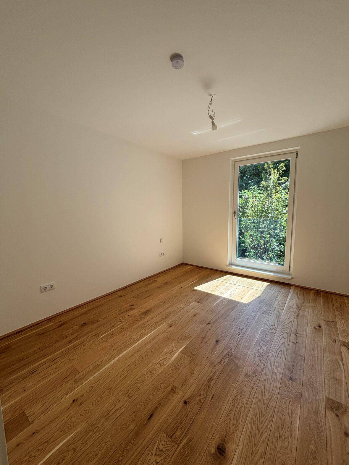Helles Schlafzimmer mit Holzboden und großem Fenster, das einen angenehmen Blick ins Grüne bietet.
