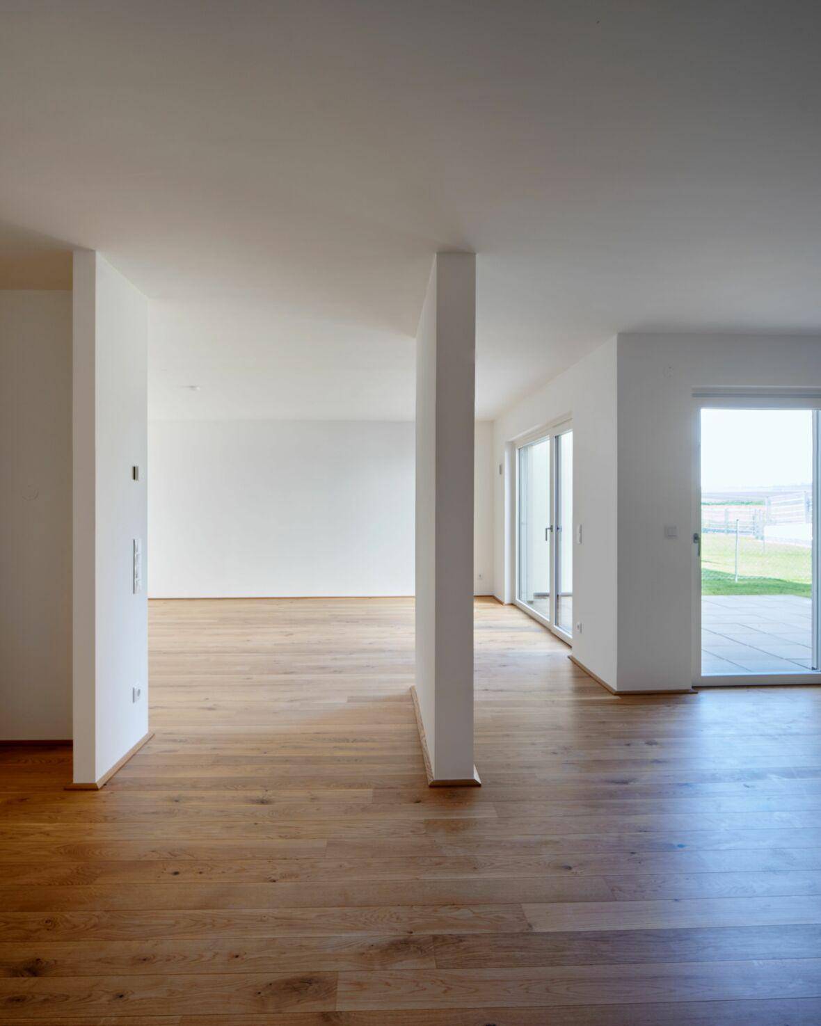 Heller, leerer Raum mit Holzboden und großen Fenstern, die viel Tageslicht hereinlassen und Zugang nach draußen bieten.