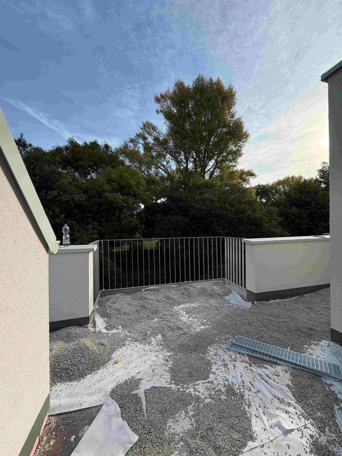 Dachterrasse im Bau mit Kiesbelag und Blick auf grüne Bäume unter blauem Himmel.