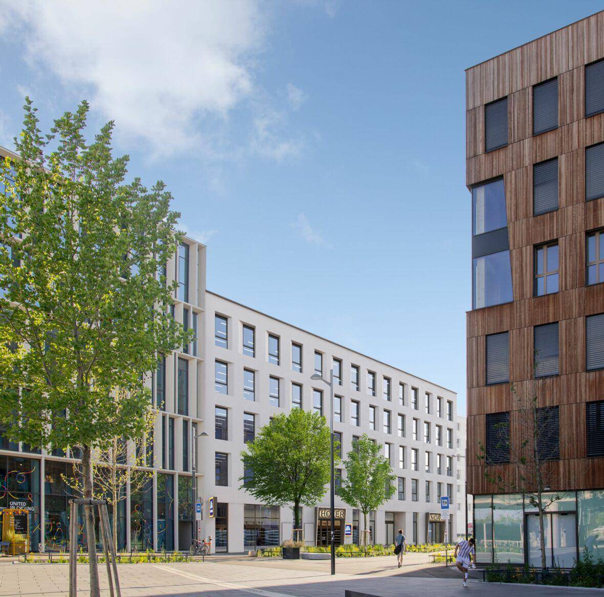 Moderne Stadtarchitektur mit verschiedenen Gebäudefassaden und begrünten öffentlichen Bereichen unter blauem Himmel.