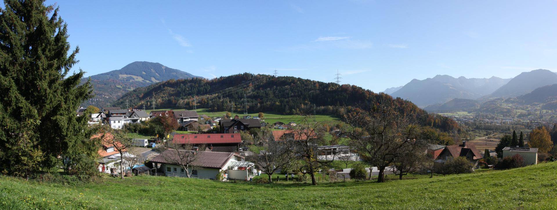 Panoramablick auf eine ländliche Siedlung mit grünen Hügeln und majestätischen Bergen im Hintergrund unter blauem Himmel.