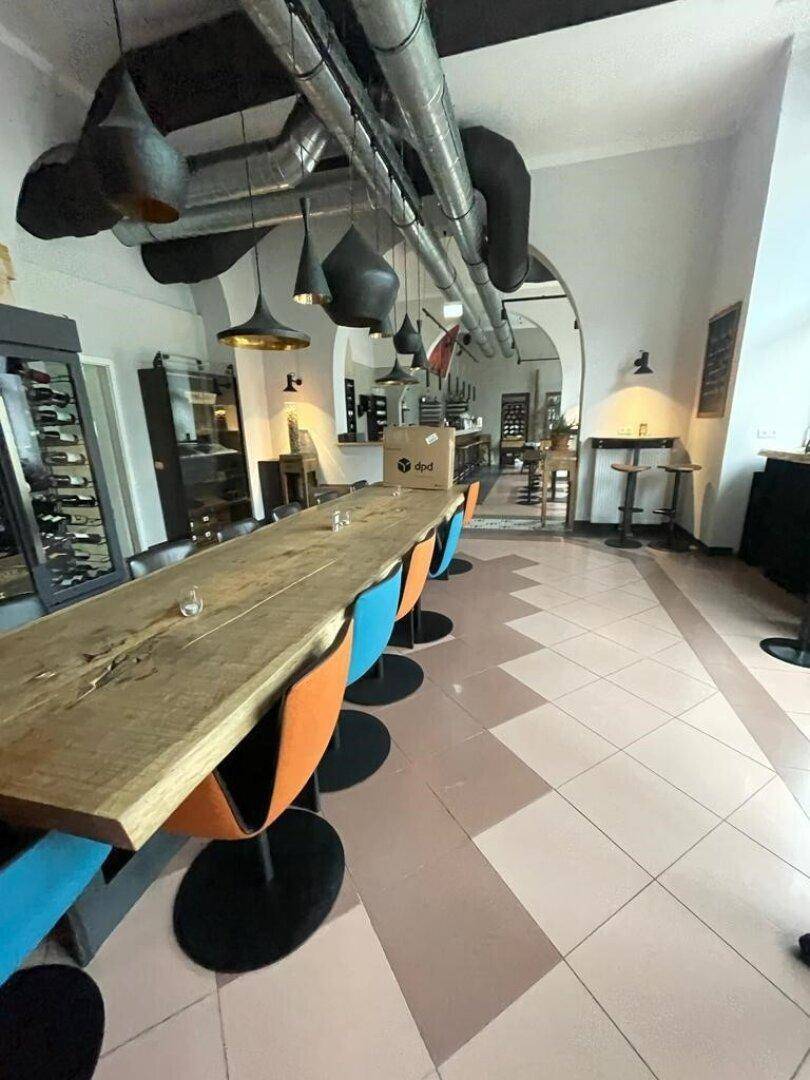 Langer Holztisch mit bunten Stühlen in einem Gastraum mit Fliesenboden und industriellen Deckenrohren.