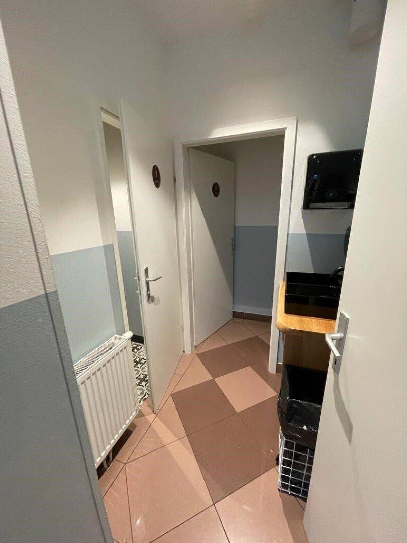 Schmaler Flur mit hellen Wänden und einem gemusterten Fliesenboden, der zu den Toiletten führt.
