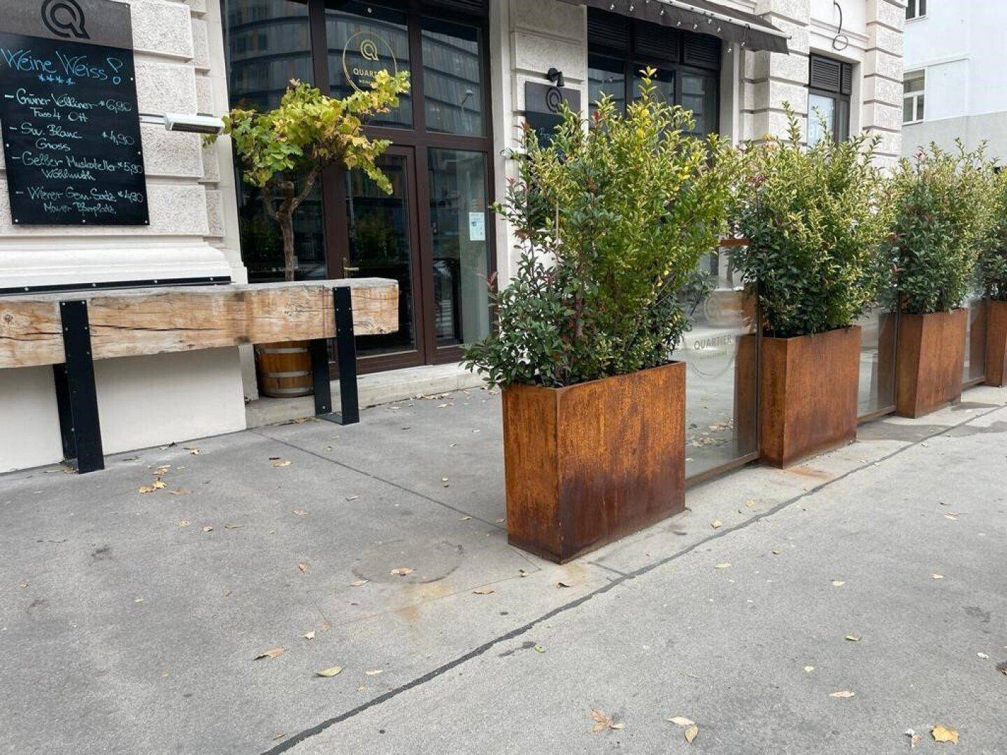 Detailansicht der Gebäudefassade mit großen Glasfenstern, Pflanzenkübeln und einer Tafel mit Angeboten auf dem Bürgersteig.
