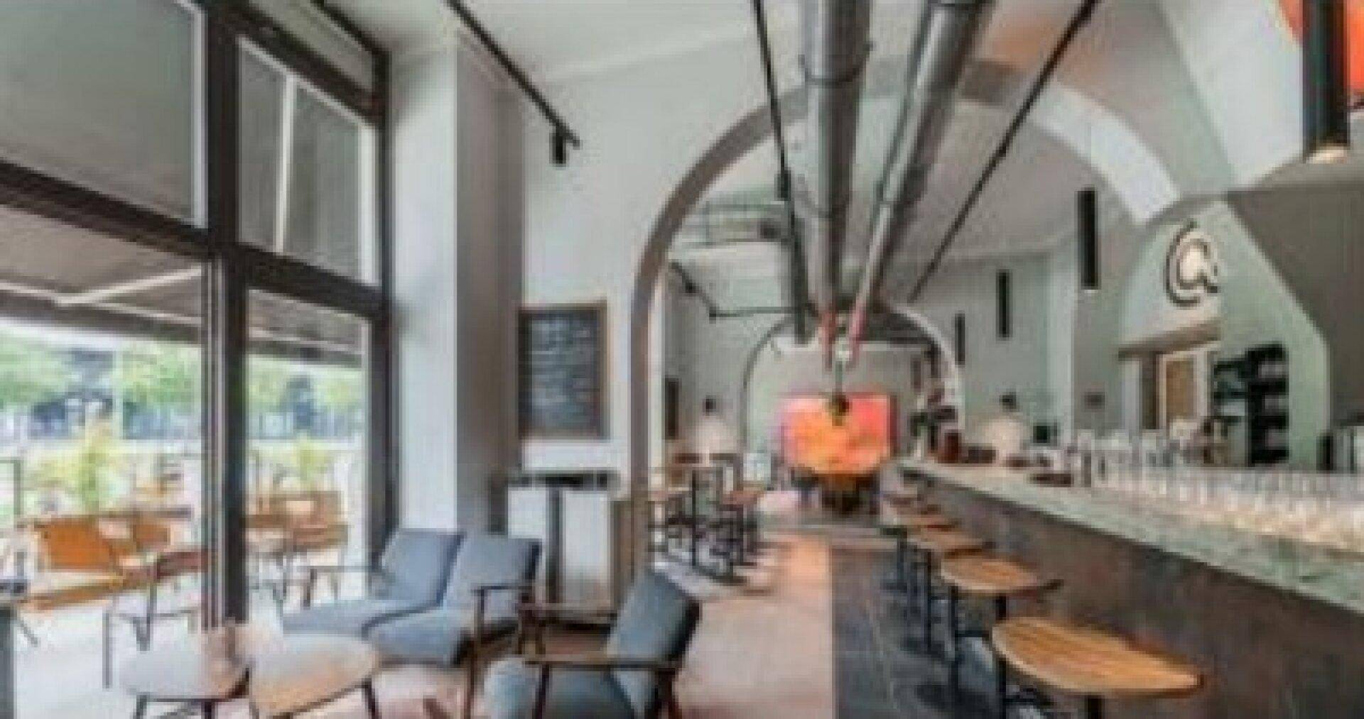 Heller Gastraum mit verschiedenen Sitzgelegenheiten, Bar und großen Fenstern, die viel Tageslicht hereinlassen.