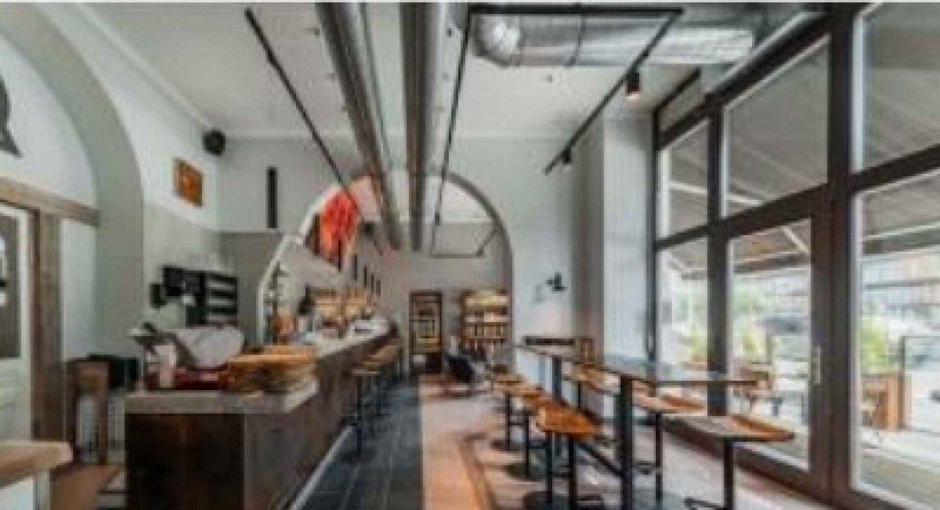 Innenansicht eines modernen Gastraums mit langer Bar, hohen Fenstern und sichtbaren industriellen Deckenrohren.