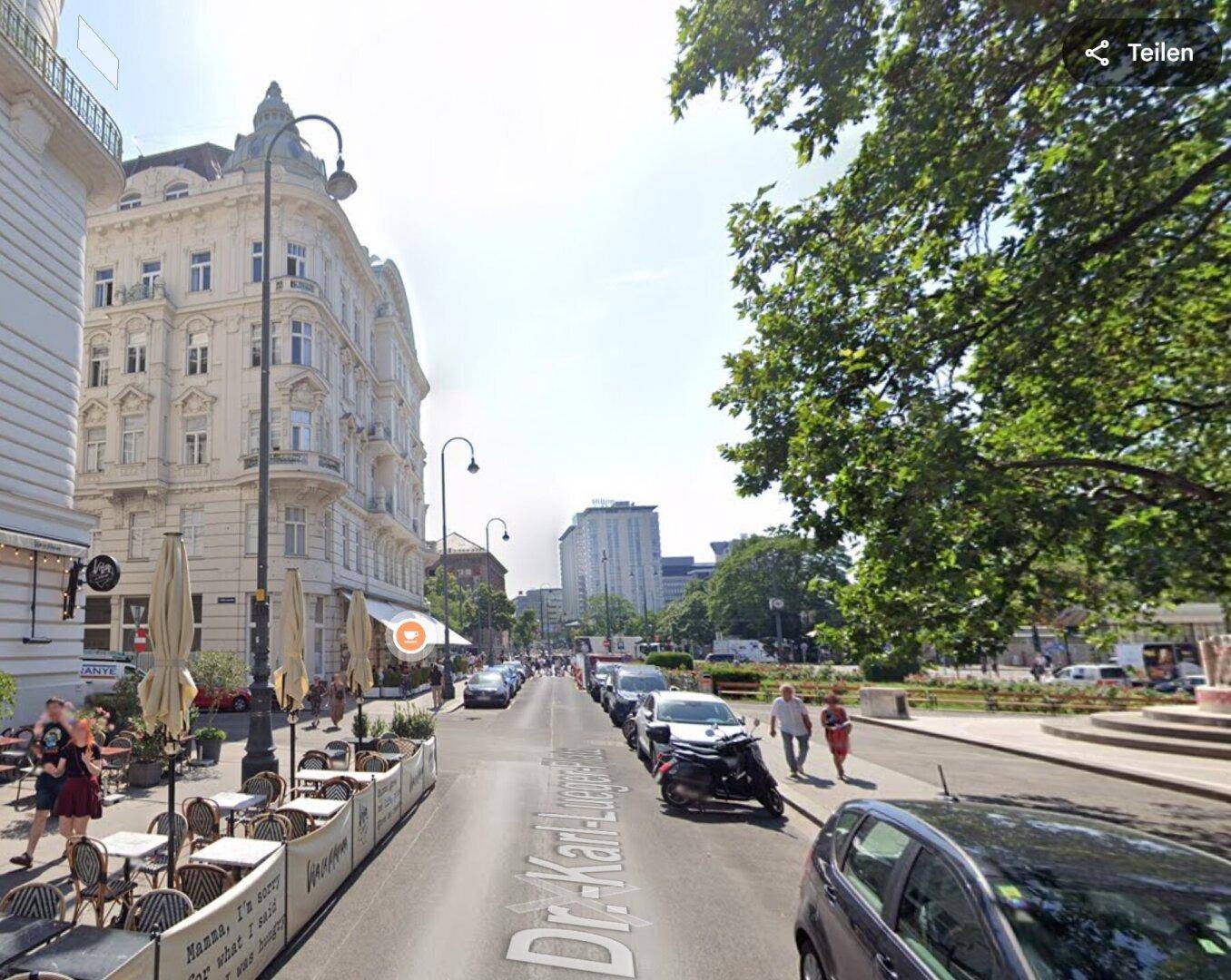 Sonniger Blick auf eine belebte Straße mit historischen Gebäuden, Straßencafés und parkenden Autos.
