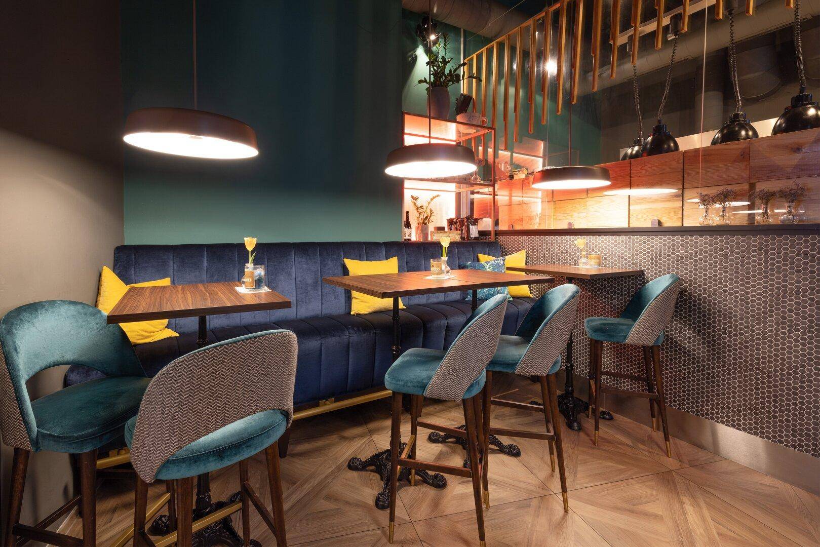 Gemütlicher Eckbereich des Restaurants mit blauer Samtbank, kleinen Holztischen und tealfarbenen Barhockern.