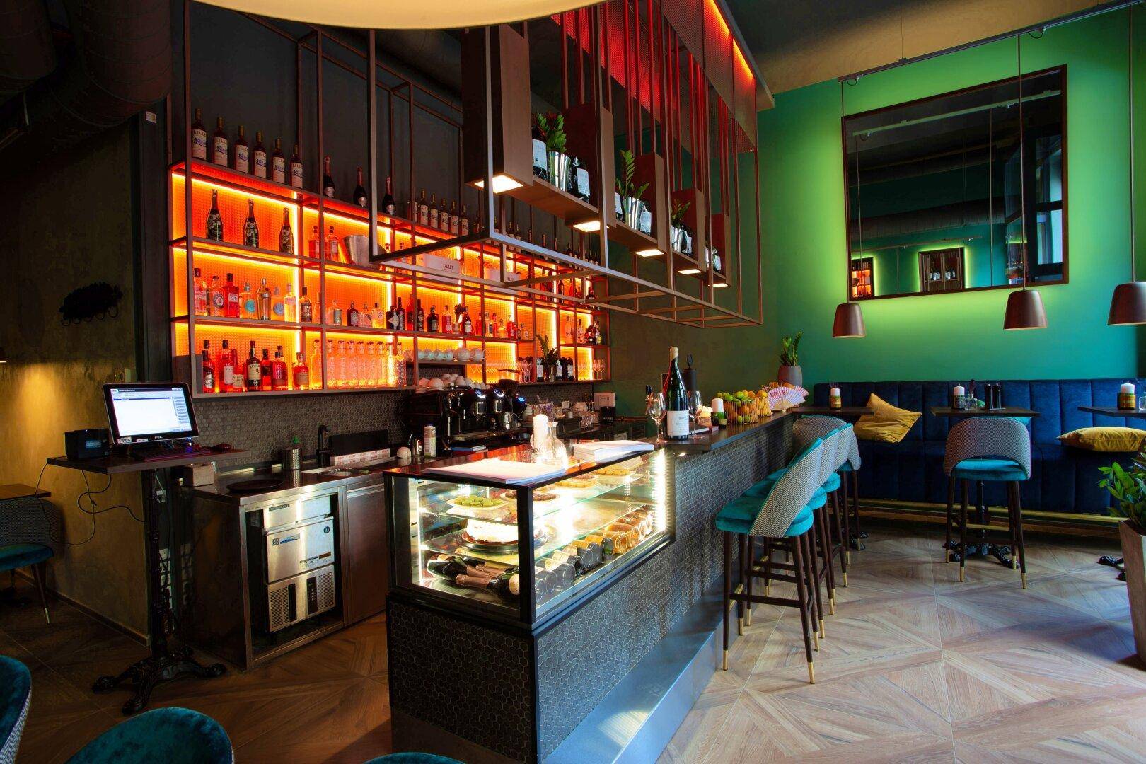 Gesamtansicht der Bar mit beleuchteten Regalen, einer Theke mit Kuchenvitrine und dem angrenzenden Sitzbereich.