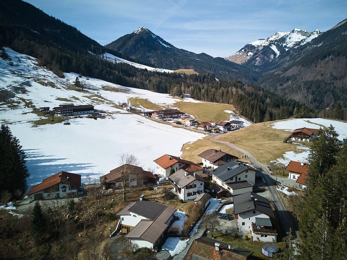 Panoramische Luftaufnahme einer ländlichen Siedlung in einer verschneiten Berglandschaft mit Wäldern.