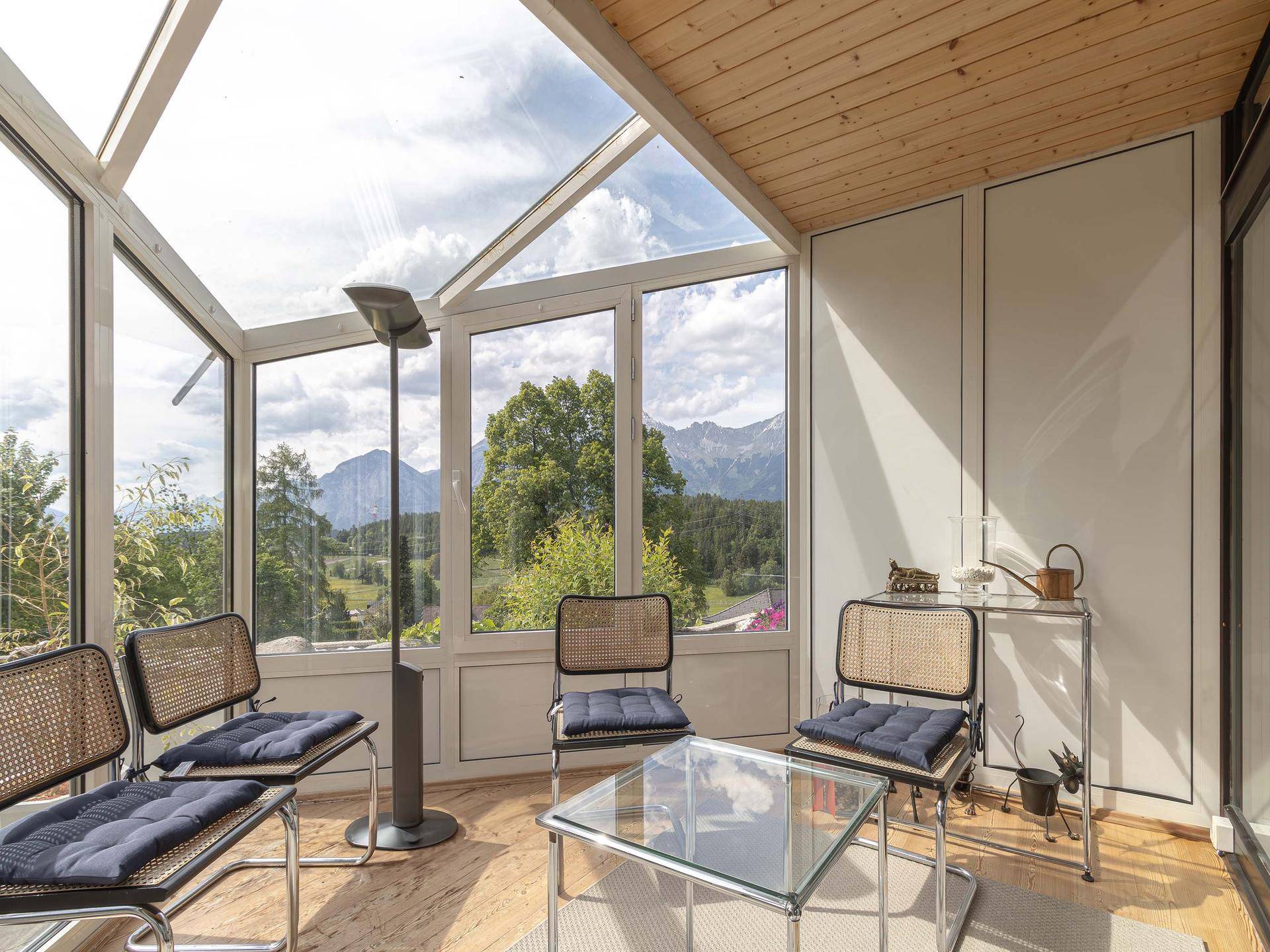 Heller Wintergarten mit Glasdach, Korbmöbeln und Panoramablick auf die Berge.
