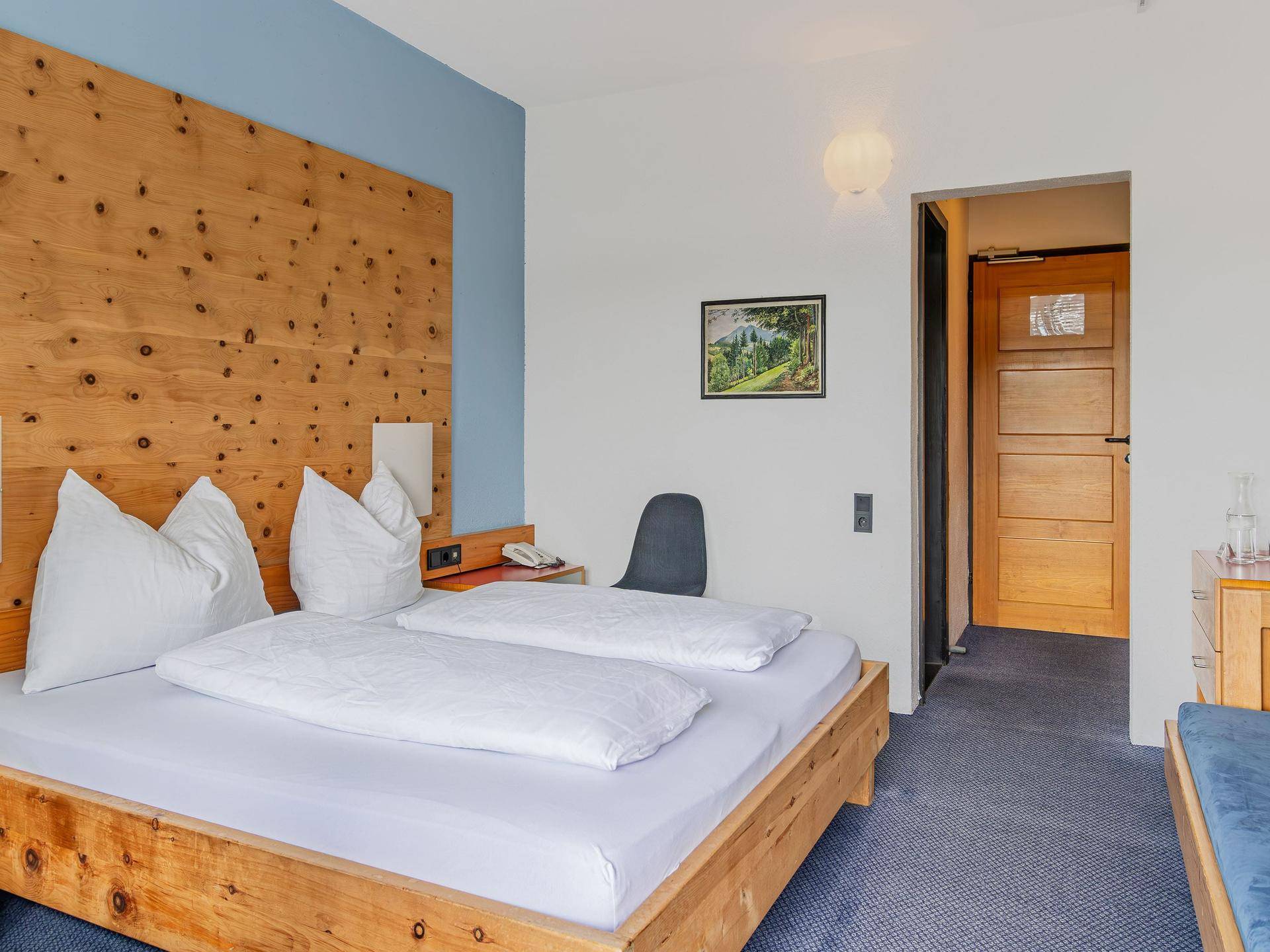 Schlafzimmer mit Holzkopfteil, blauem Teppichboden und einfacher, funktionaler Einrichtung.