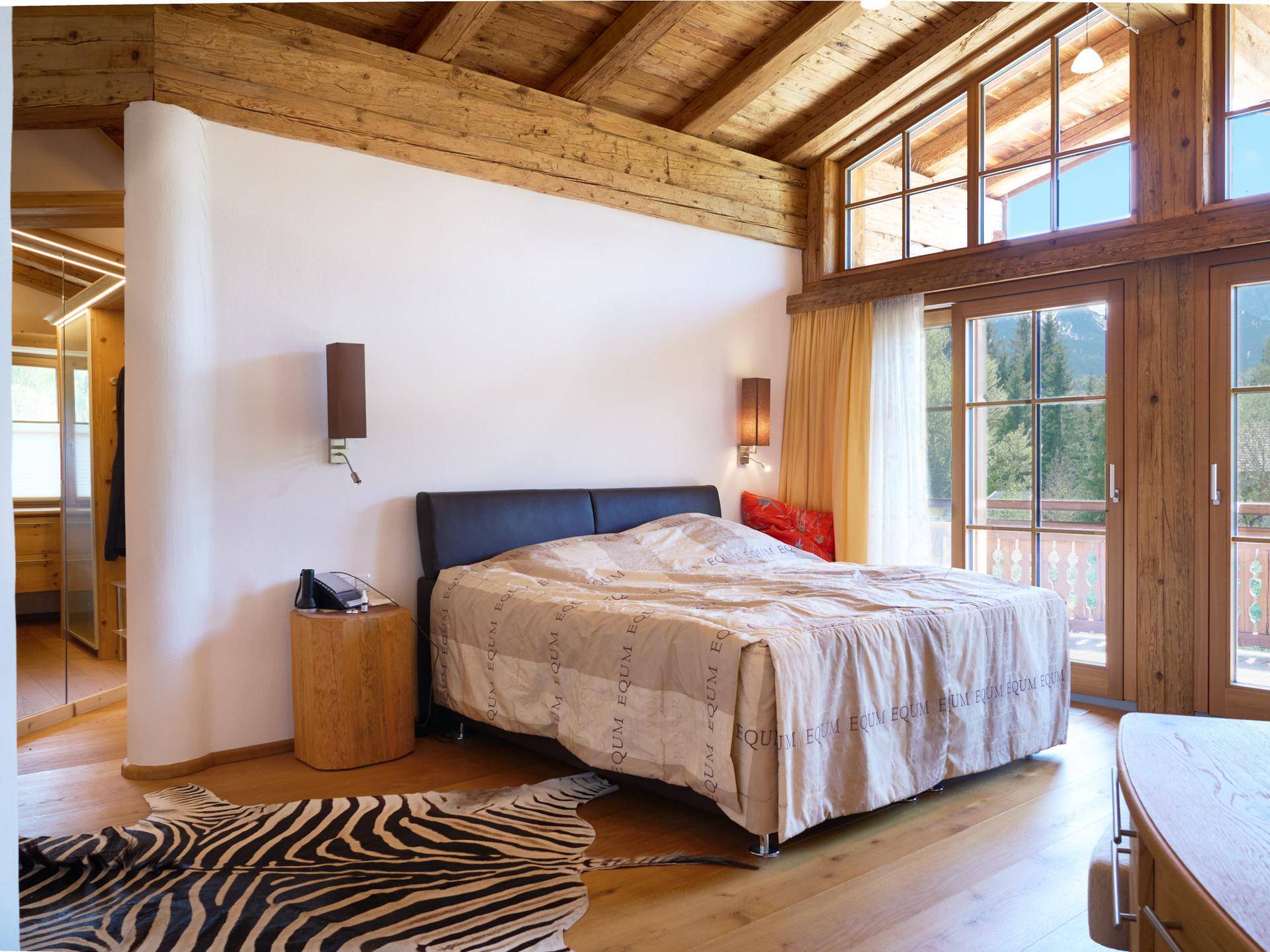 Schlafzimmer im alpinen Stil mit Holzbalken, großen Fenstern und Zebra-Teppich.